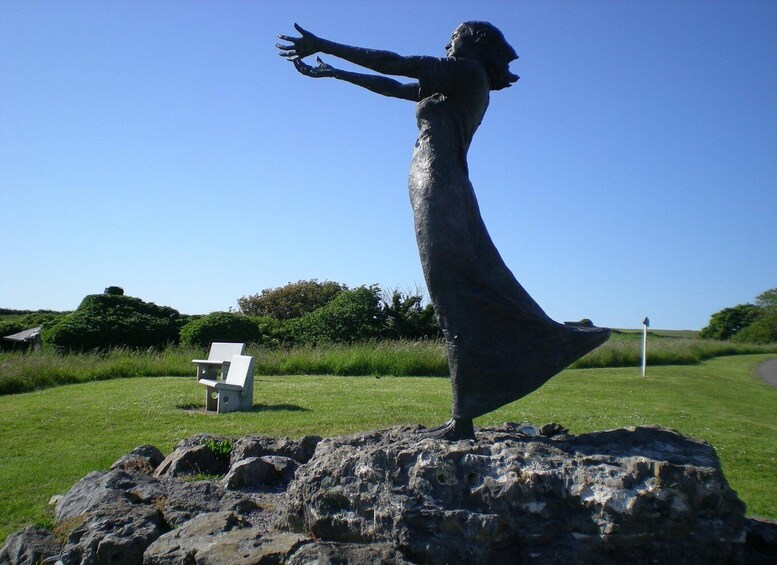 A statue of a woman in a flowing dress standing on a rock with a grassy field and trees in the background. 4