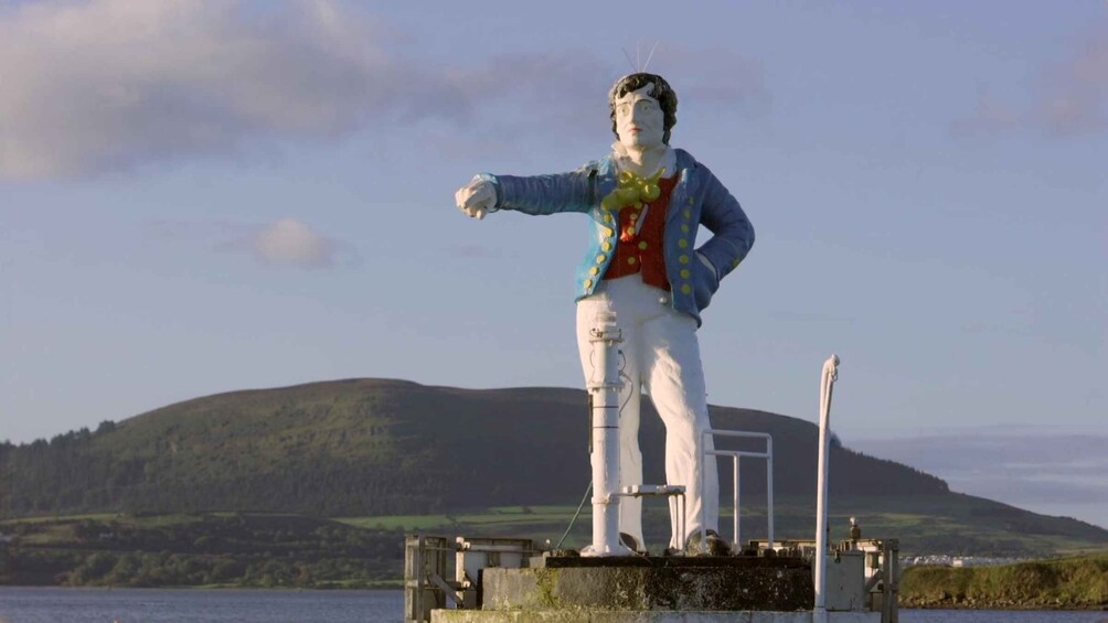 A statue of a man in a blue jacket and white pants, standing on a platform by a body of water with hills in the background. 5