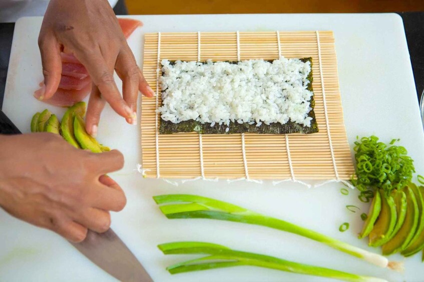Hands preparing sushi with rice, avocado, and green onions on a cutting board. 3