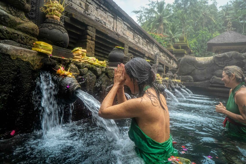 A person in a green garment performing a ritual by a water feature with a stone structure and a thatched roof in the background. 2