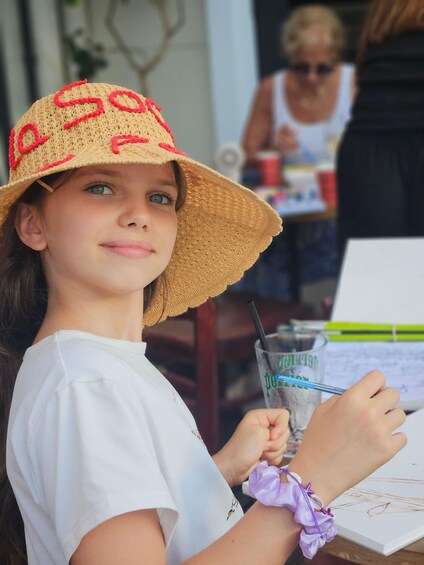A child wearing a straw hat with red embroidery, holding a drink with a straw, and sitting at a table with a notebook. 5