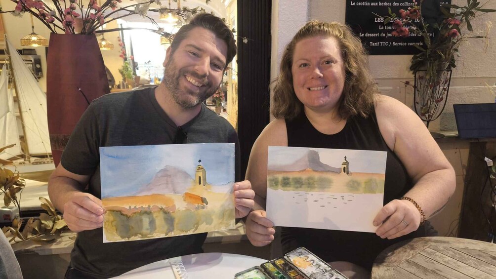 Two people holding watercolor paintings of a coastal landscape with a lighthouse. 2