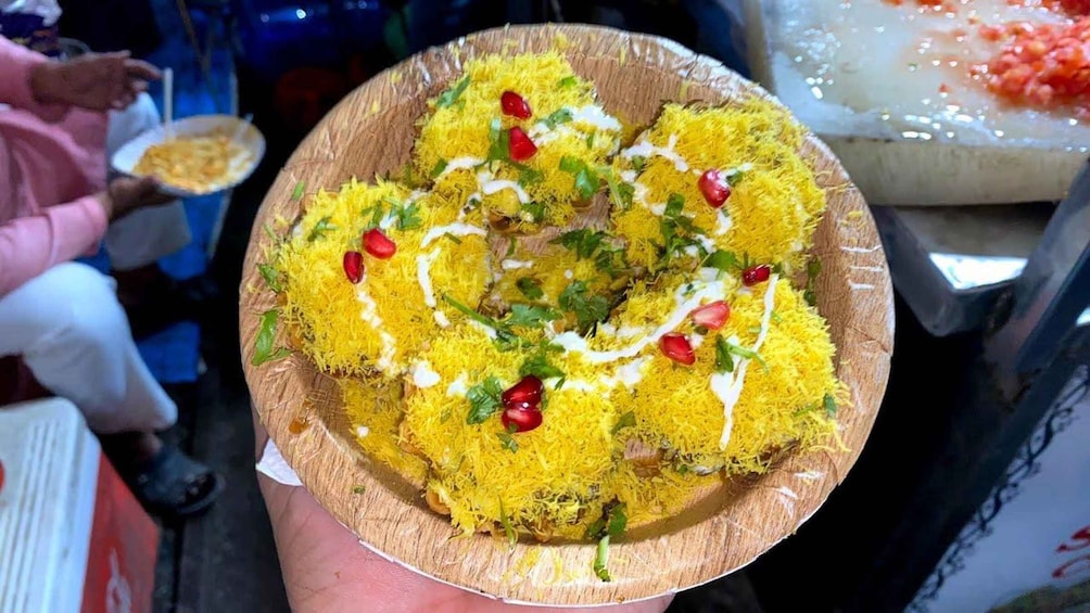 A person holding a paper plate with a food item topped with yellow shredded toppings, red pomegranate seeds, and green herbs. 4