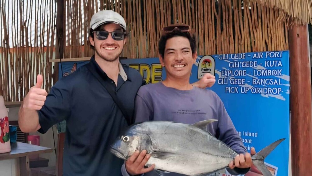 Two people holding a fish, with a sign in the background advertising activities like air port, kuta, and pick up or drop. 5