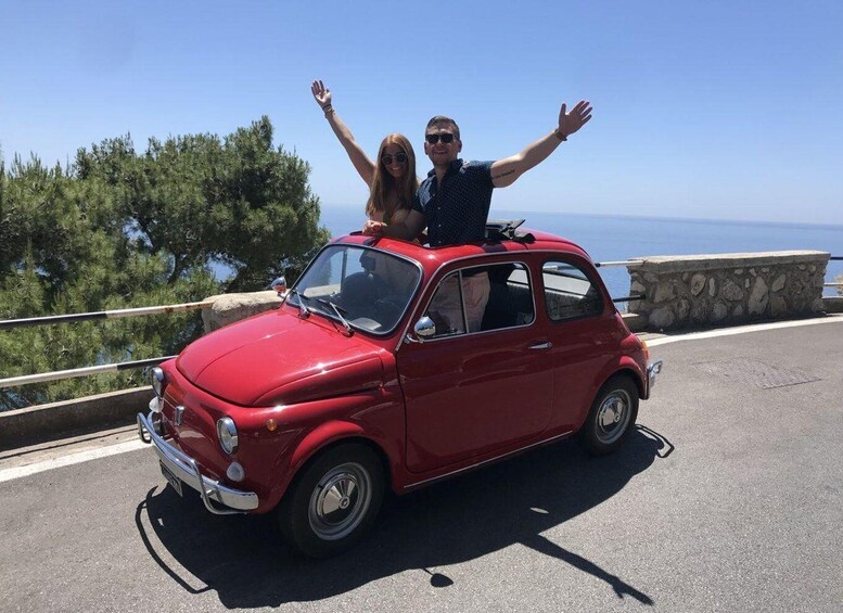 A red vintage car with two people on the roof, parked on a coastal road with a stone wall and trees in the background. 1