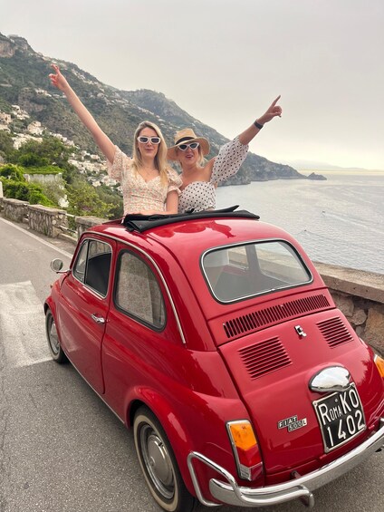 Two people on a red vintage car with a license plate 'ROMA 1402' on a coastal road with mountains in the background. 4