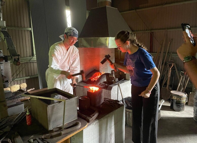 Two people working with a forge, one hammering a piece of metal, in a workshop with tools and a large chimney. 1