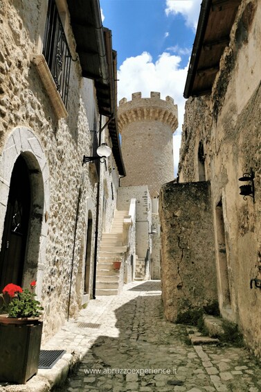 A narrow cobblestone street flanked by historic stone buildings with a tower in the distance. 1