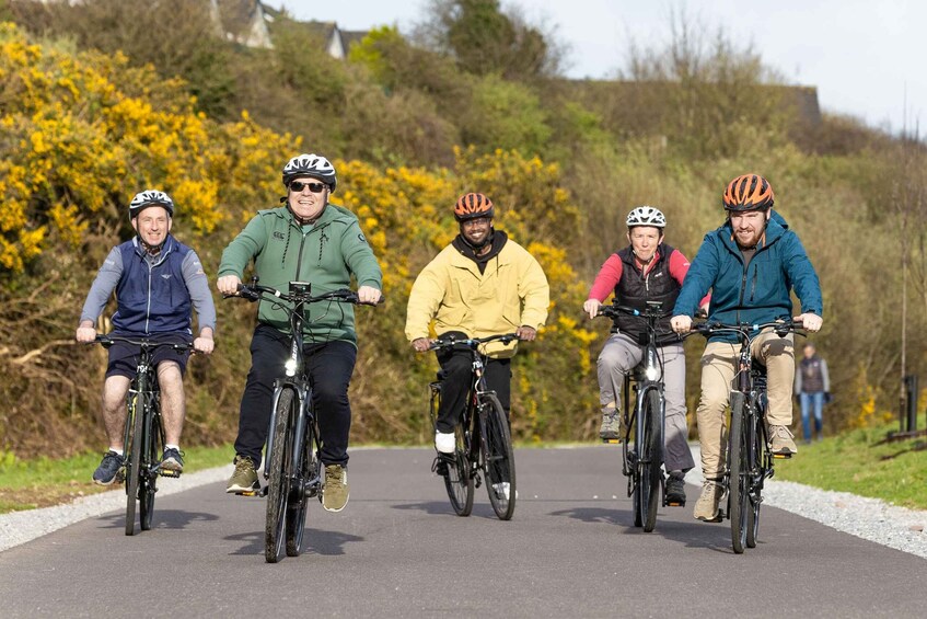 A group of cyclists on a paved path with yellow flowering bushes in the background. 3
