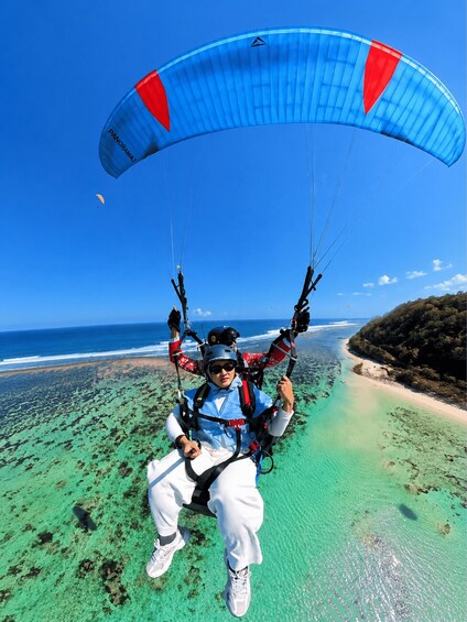 A person paragliding over a beach with clear turquoise water and a distant shoreline. 1