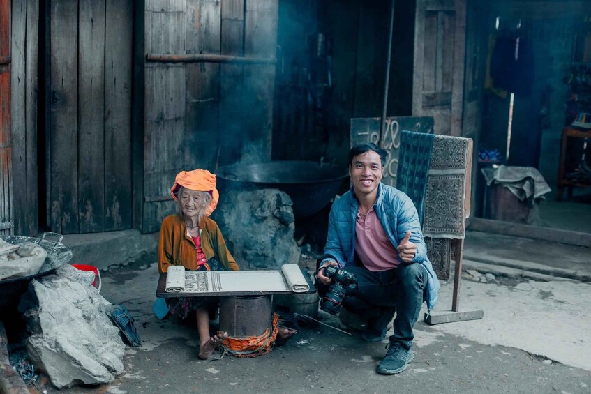 An elderly person sitting next to a large metal pot emitting steam, with a young person crouching nearby holding a camera. 3