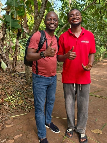 Two people standing outdoors, one giving a thumbs-up, in a natural setting with trees and foliage. 4