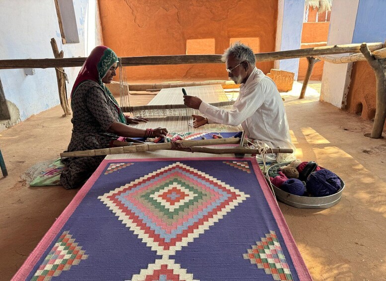Two people weaving on a loom with a large, colorful woven mat in front of them. 3