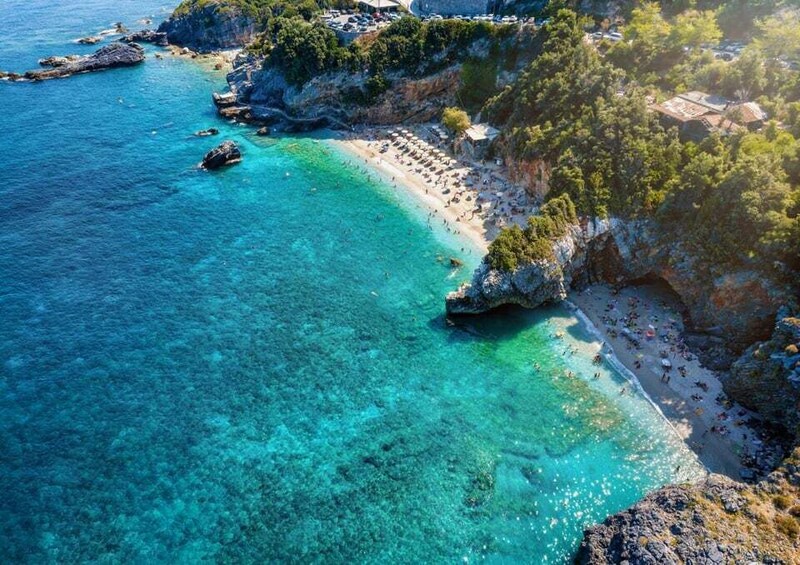 A coastal landscape with clear turquoise waters, rocky cliffs, and a beach populated with people. 5