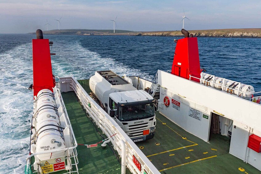 A ferry with a white truck and a red funnel, traveling on a body of water with wind turbines in the background. 2