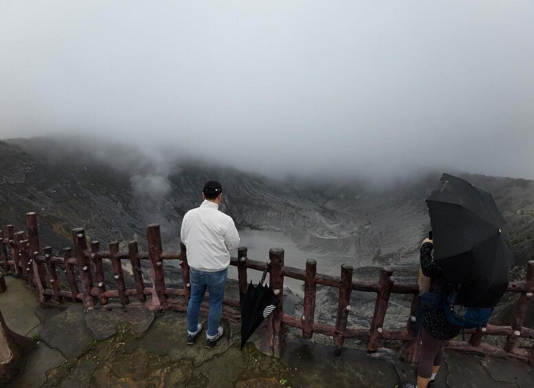 Two people standing by a wooden fence overlooking a volcanic crater with steam rising from the crater's edge. 5