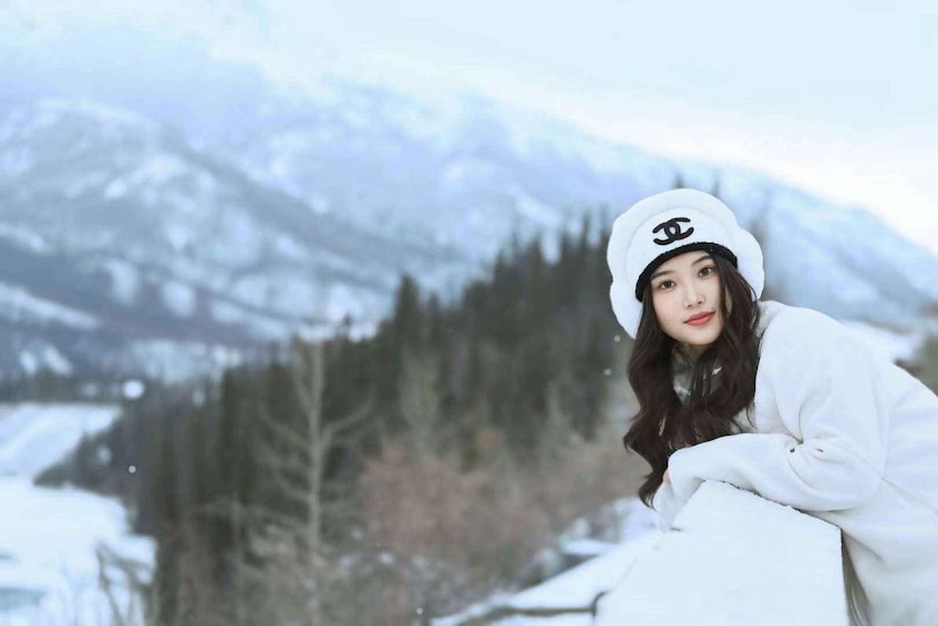 A person in a white jacket and hat with a visible logo, sitting outdoors with snow-covered trees and mountains in the background. 1