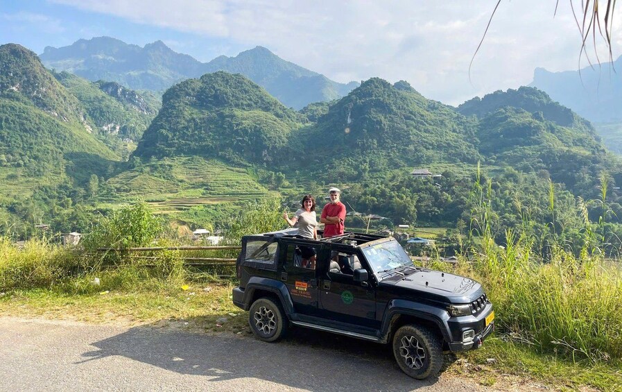 A black off-road vehicle parked on a road with two people standing on the roof, surrounded by lush green hills and mountains in the background. 1