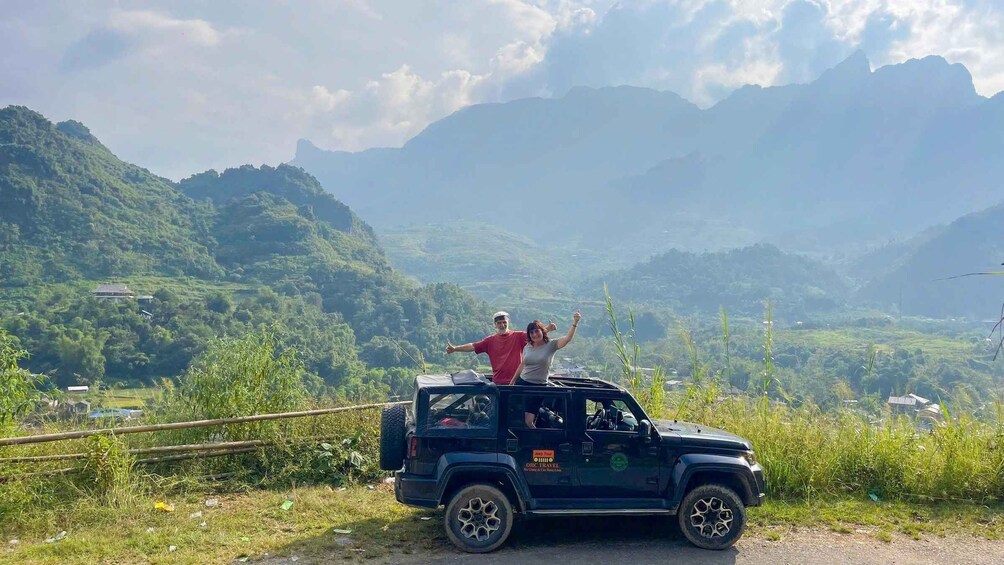 A black SUV parked on a road with two people standing on the roof, surrounded by lush green hills and mountains in the background. 2