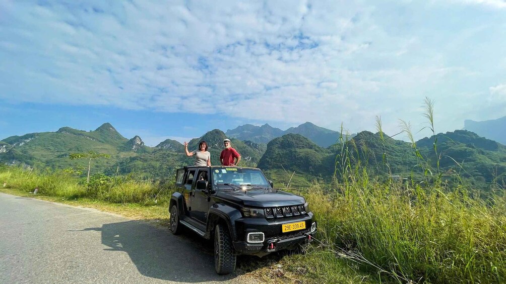 A black SUV on a road with two people standing on the roof, surrounded by lush green hills and a clear sky. 3