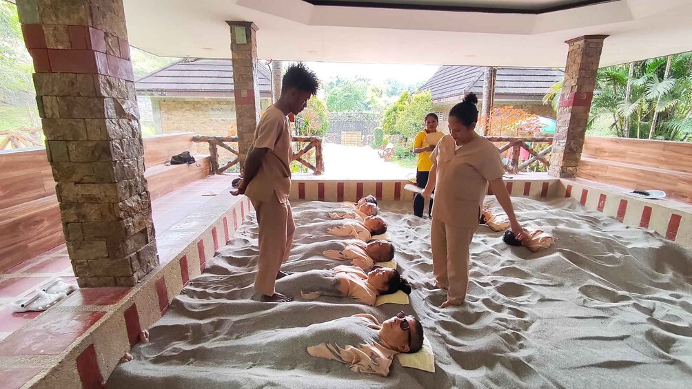 A group of people lying on sand in a covered area with stone pillars and a wooden bench in the background. 1