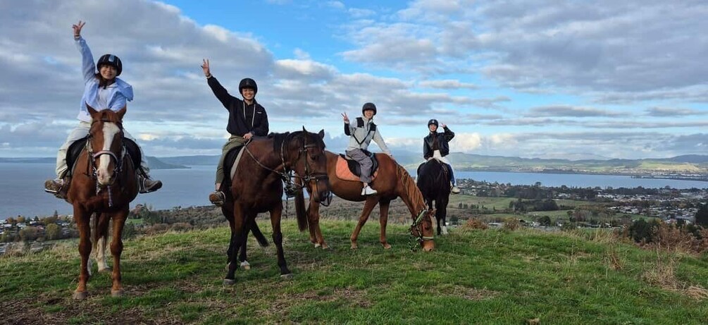 Four people riding horses on a grassy hill with a lake and distant town in the background. 1