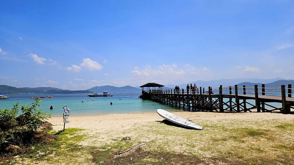 A beach with a wooden pier extending into the water, a surfboard on the sand, and boats in the distance. 3