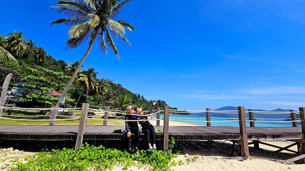 A wooden pier with two people sitting, overlooking a beach with palm trees and clear blue water. 1