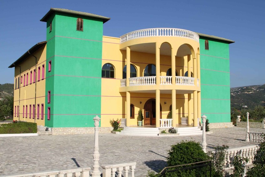 A large, two-story building with a green and yellow facade, a balcony with white balustrades, and a stone-paved courtyard. 4