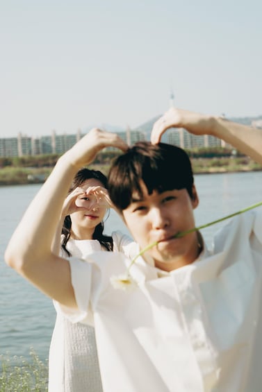 Two people making a heart shape with their hands, standing by a body of water with a bridge in the background. 1