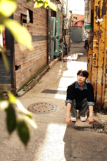 A person crouching on a concrete surface in an alleyway with wooden buildings and a metal grate. 4