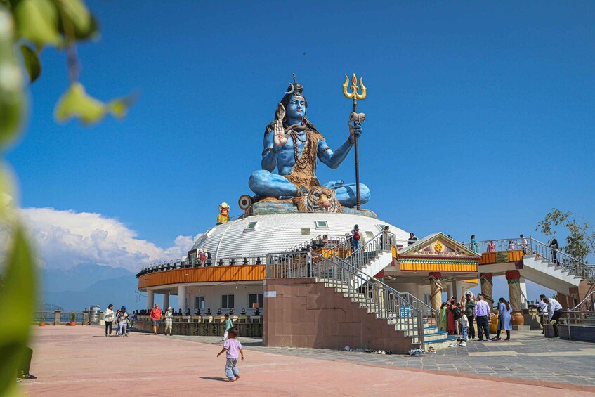 A large statue of a deity with a trident, seated on a platform with a dome, surrounded by visitors and a building with stairs. 4