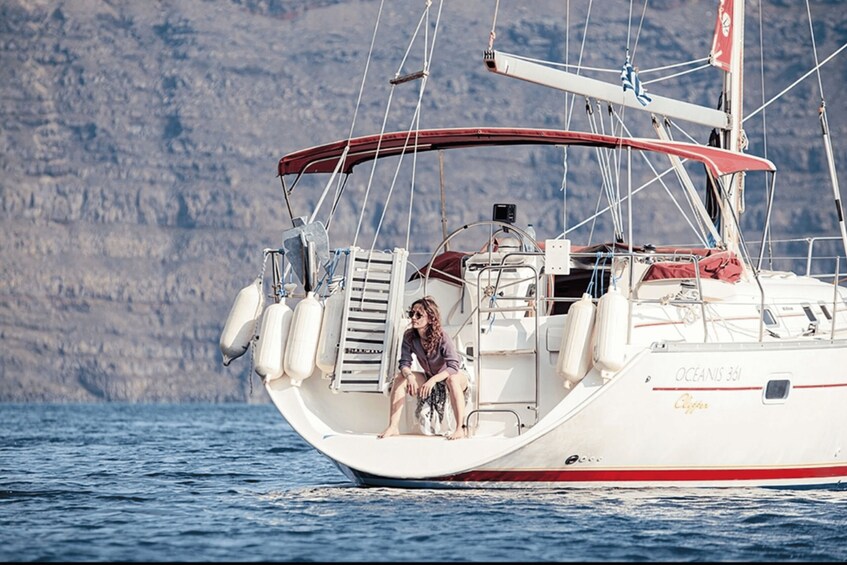 A woman sitting on the deck of a sailboat named 'OCEANS 361' with a mountainous coastline in the background. 4