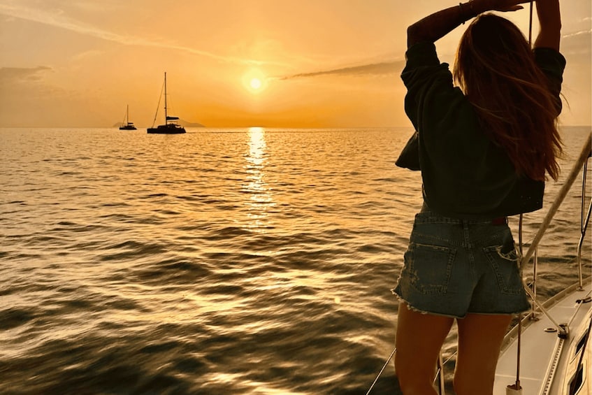 A person standing on a boat, facing the sunset over the ocean with sailboats in the distance. 1