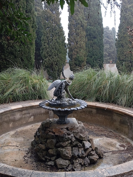 A stone fountain with a statue of a figure in a dynamic pose, surrounded by lush greenery and tall trees. 4