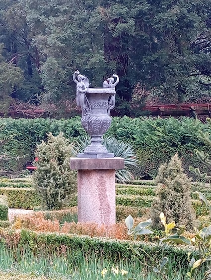 A stone urn with figures, surrounded by a manicured garden with various shrubs and flowers. 2