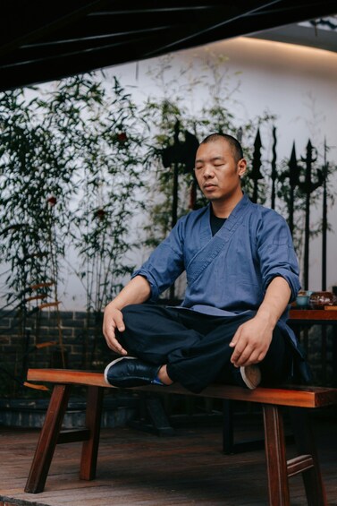 A person sitting on a wooden bench outdoors with bamboo plants and a fence in the background. 4