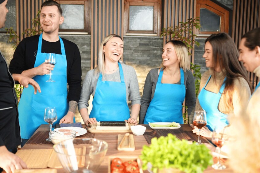 A group of people wearing aprons, standing around a table with food and wine. 2