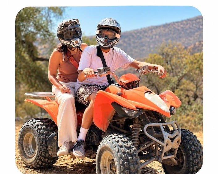 Two people on an orange ATV in a desert landscape with sparse vegetation and hills in the background. 1