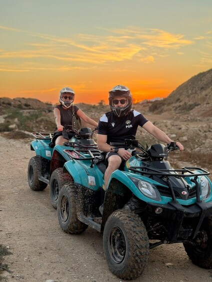 Two people riding a teal ATV on a dirt path with a sunset in the background. 2