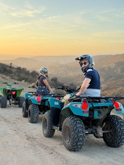 Two people riding ATVs on a dirt road with a mountainous landscape in the background. 3
