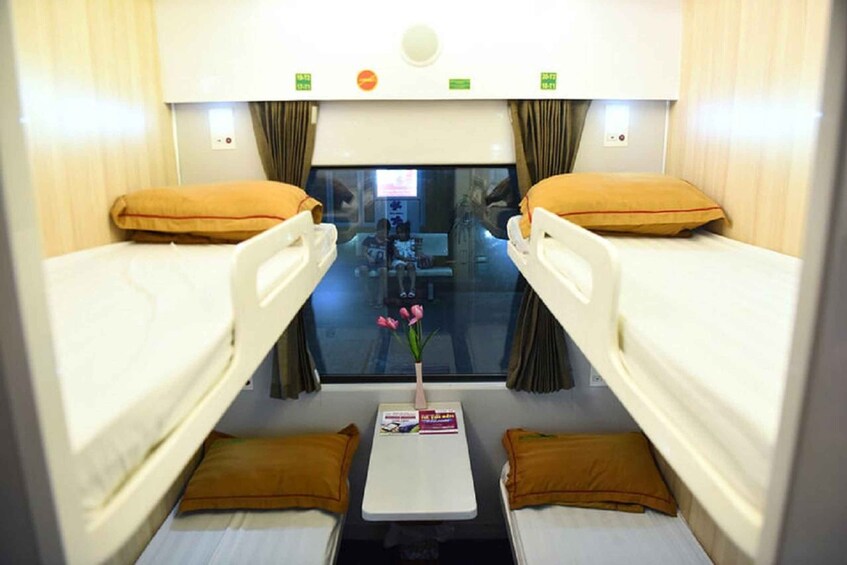A compact interior with two beds, a small table with a magazine, and a window with curtains. 2