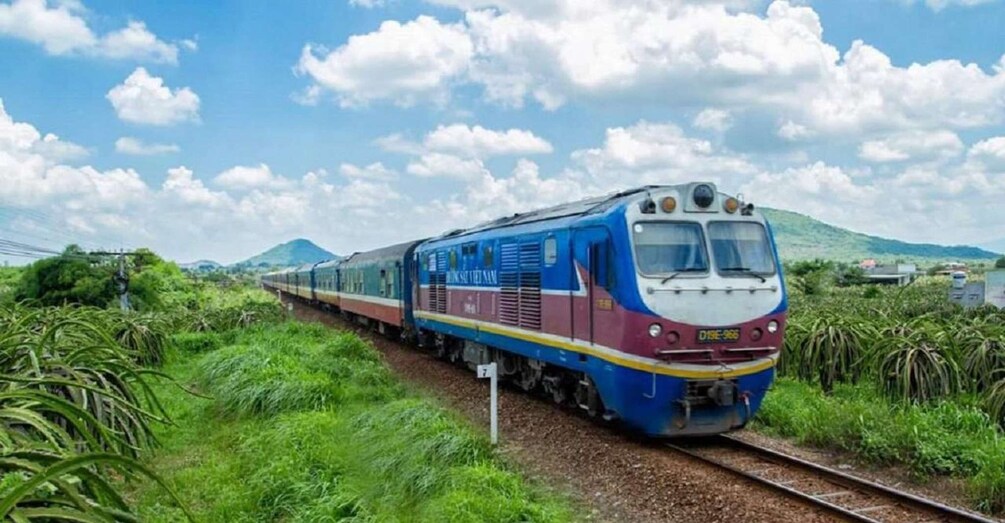 A blue and red train traveling through a lush green landscape with mountains in the background. 1