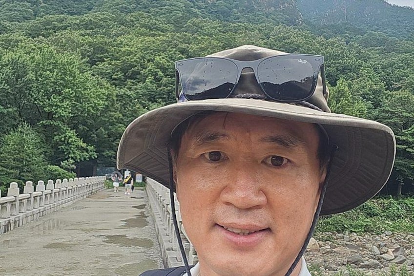 A person wearing a hat and sunglasses, standing on a bridge with a forested landscape in the background. 2