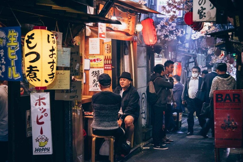 A crowded street with people standing and sitting, signs in Japanese, and a red sign that says 'BAR'. 3