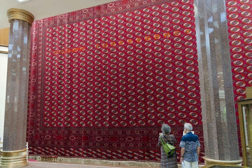 A large red patterned carpet with geometric designs, displayed between two columns in a building. 4