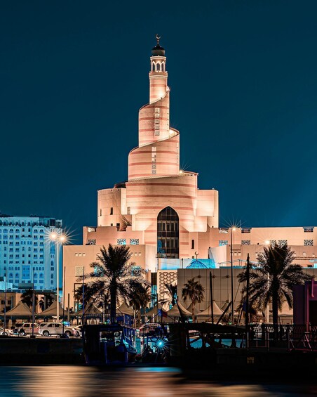A large, illuminated building with a spiral minaret, palm trees, and a body of water in the foreground. 1