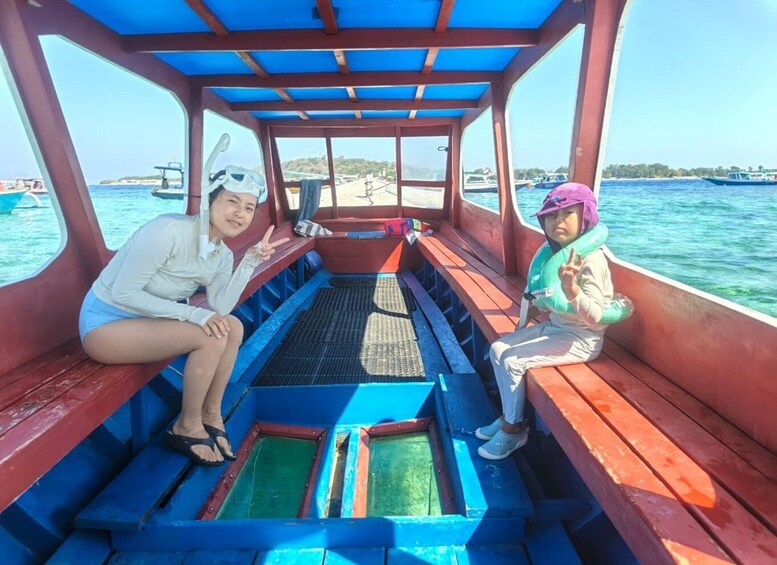 A boat with two people sitting on the deck, a blue and red interior, and a view of the sea and other boats in the background. 1