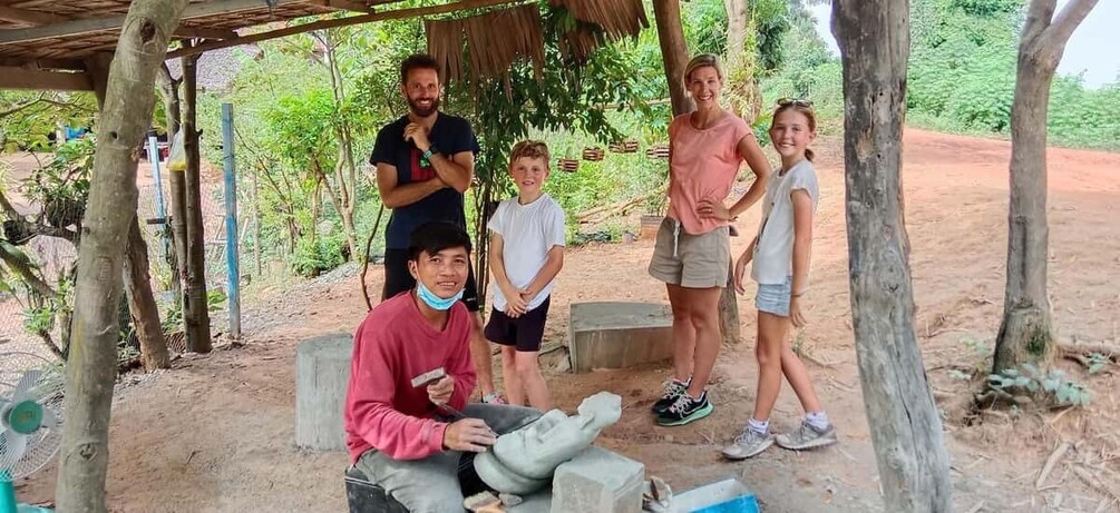 A group of people outdoors under a wooden shelter, with trees and a dirt ground. 5