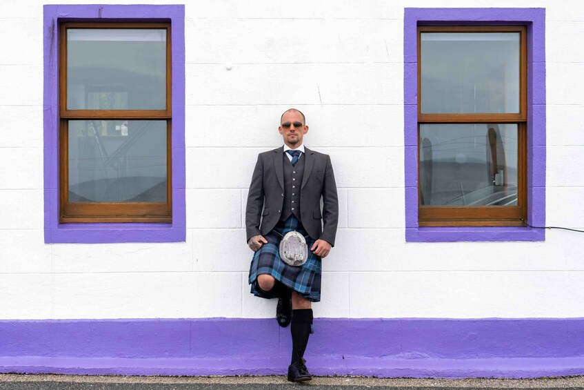 A person in a kilt and suit standing between two windows with purple frames. 3